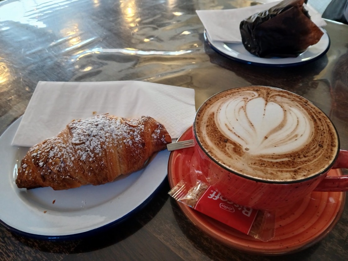 An almond croissant dusted with icing sugar beside a coffee in a red speckled cup